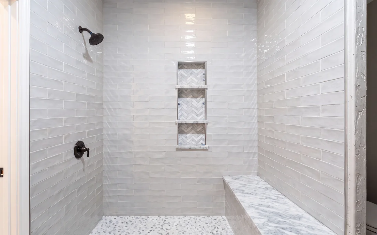 White subway-tile walk-in shower with herringbone mosaic niches, marble bench, and pebble floor