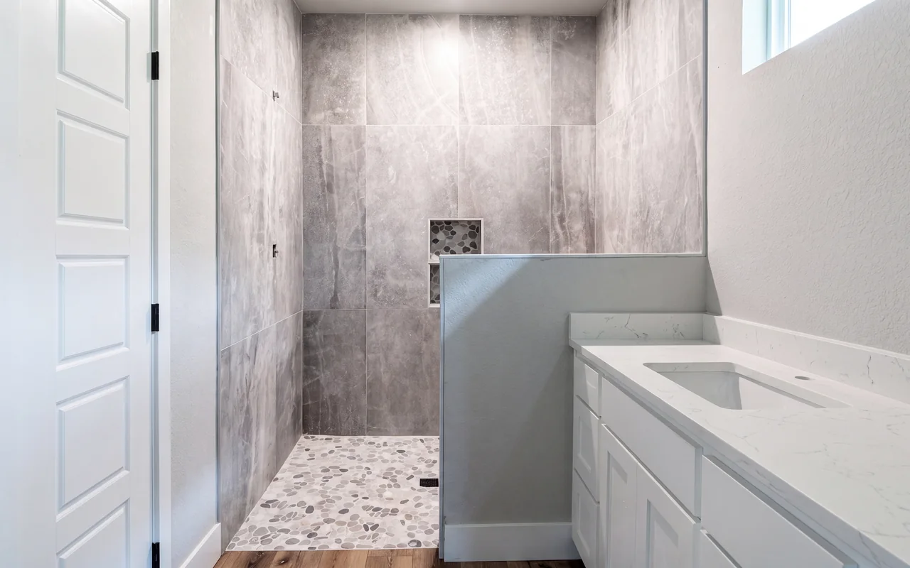 Walk-in shower with large-format stone-look tile and pebble floor beside a white quartz vanity